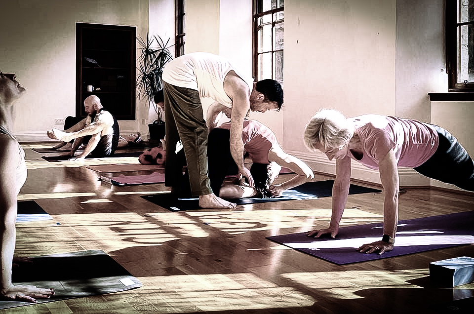 Man adjusting woman in kapotasana with other people practicing Ashtanga