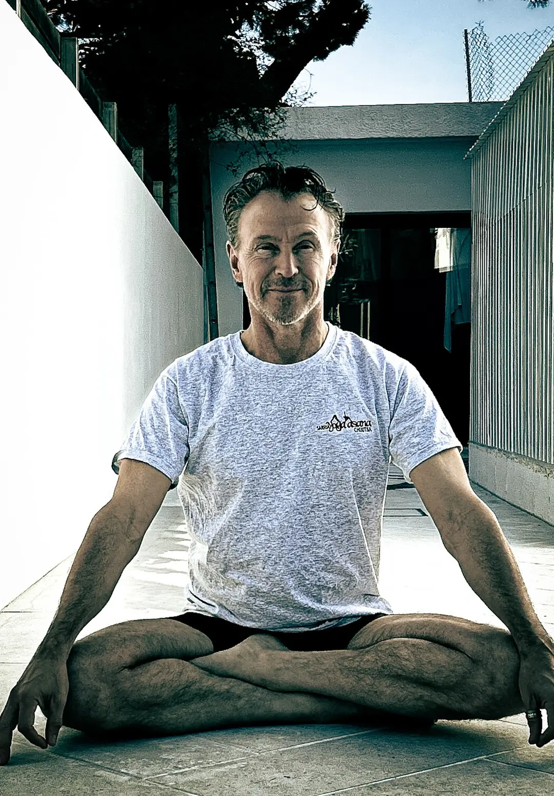Santiago pinto practicing Ashtanga Vinyasa Yoga cross-legged on a sidewalk, smiling at the camera.